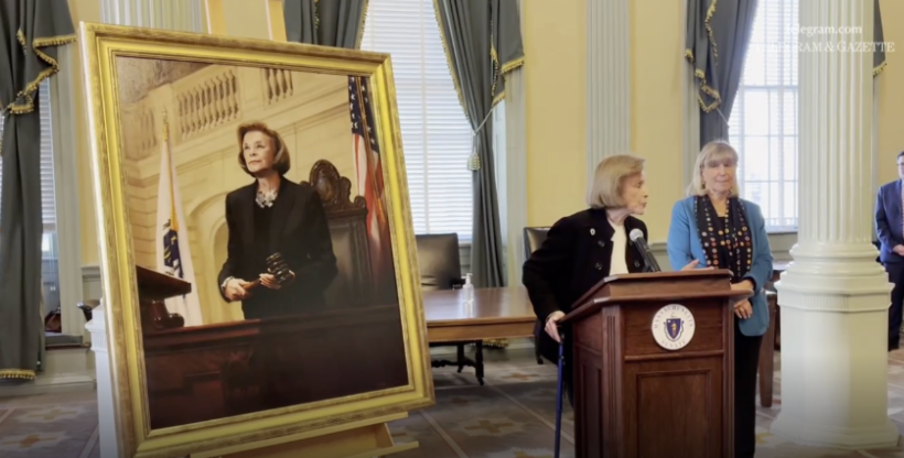 A large gold-framed oil portrait of Harriette Chandler stands on an easel in a State House chamber. Chandler stands at a lectern beside it with another woman during the unveiling ceremony.