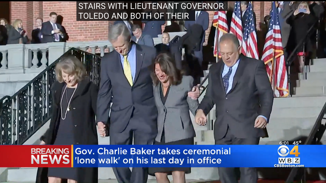 Gov. Charlie Baker and Lt. Gov. Karyn Polito walk down the State House steps with their spouses, past rows of American flags, in a televised ceremony. A CBS Boston chyron reads: Gov. Charlie Baker takes ceremonial lone walk on his last day in office.